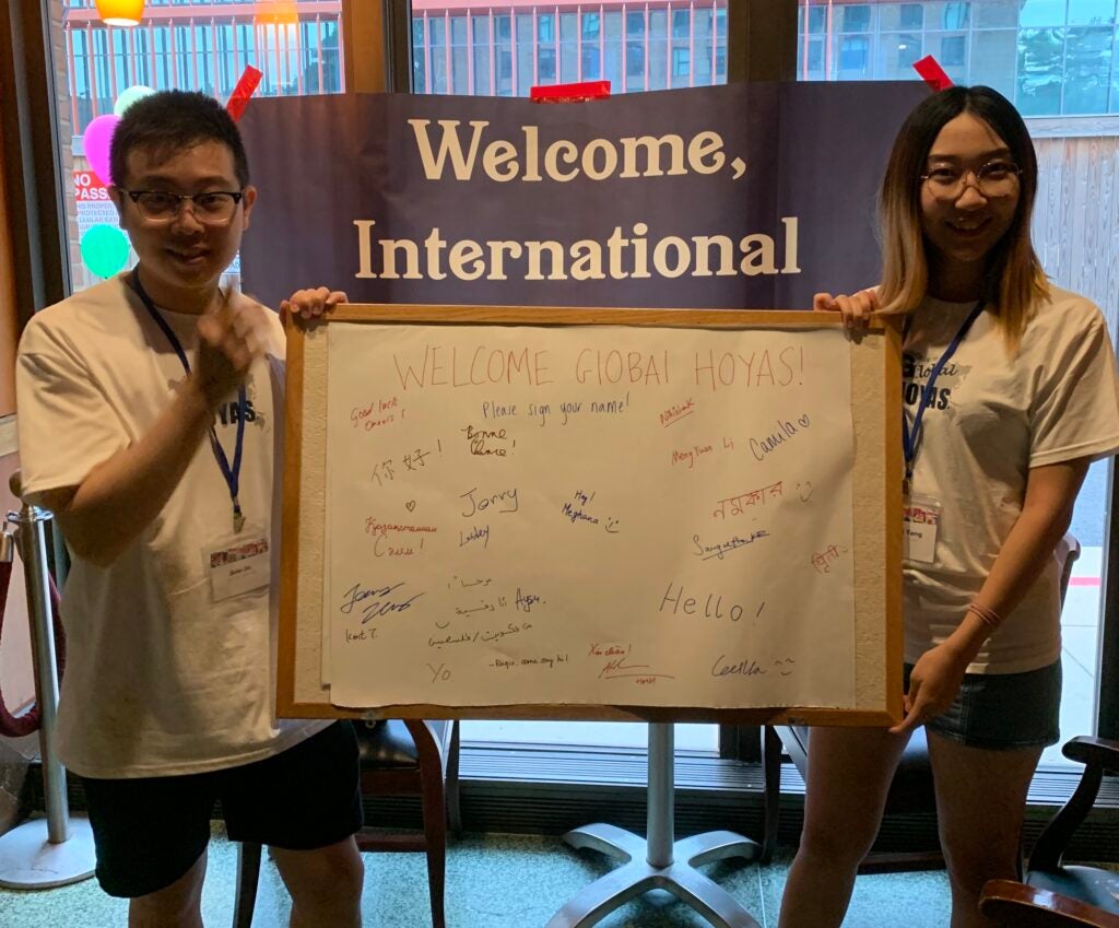Two international students hold a white poster with names of new international students written on it. Behind them is a sign that reads "Welcome, International"
