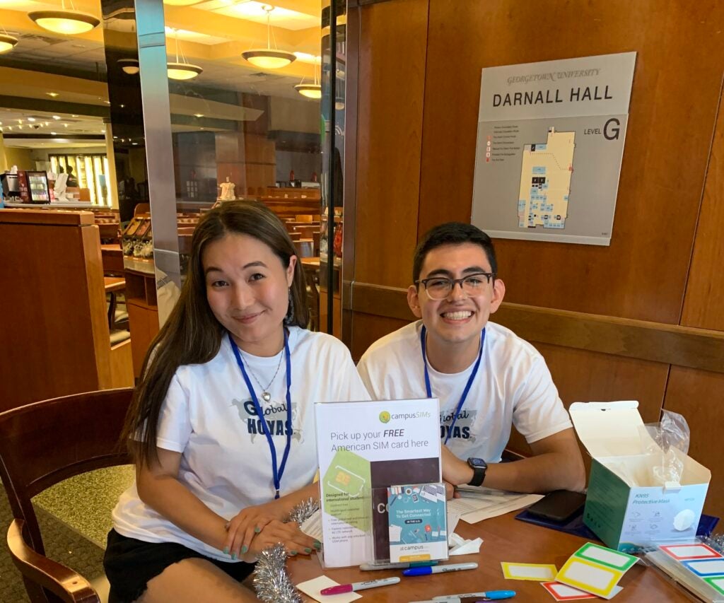 Two international students sit at a table smiling behind a sign that advertises a free American SIM card